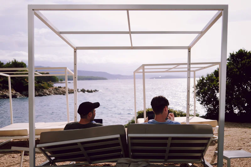 Two men sit in patio chairs under a white metal canopy overlooking the ocean and coastline.