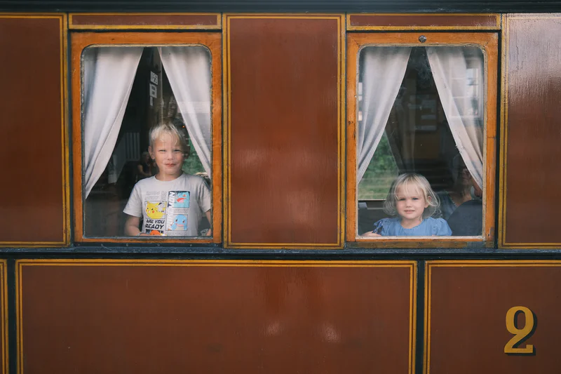 Two children sit behind the windows of a brown vintage train car with number “2” on the side.