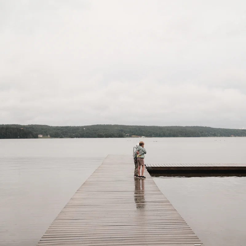 Two children stands on a wooden dock extending into a calm lake, with forests and clouds in the background.