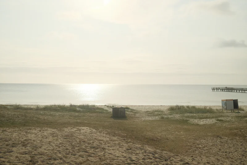 Distant calm ocean under a cloudy sky with sandy dunes, a small trash bin, and a sign near the right edge.