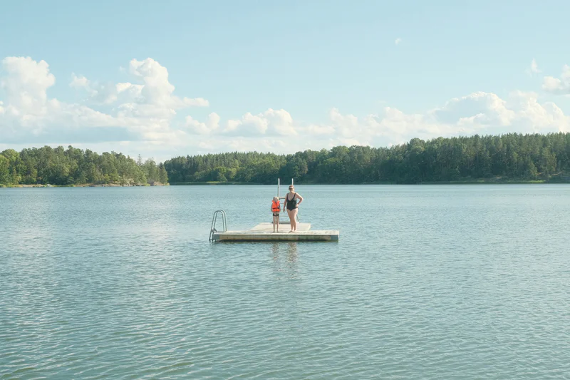 Two people stand on a small floating platform in a calm lake with trees and clouds in the background.