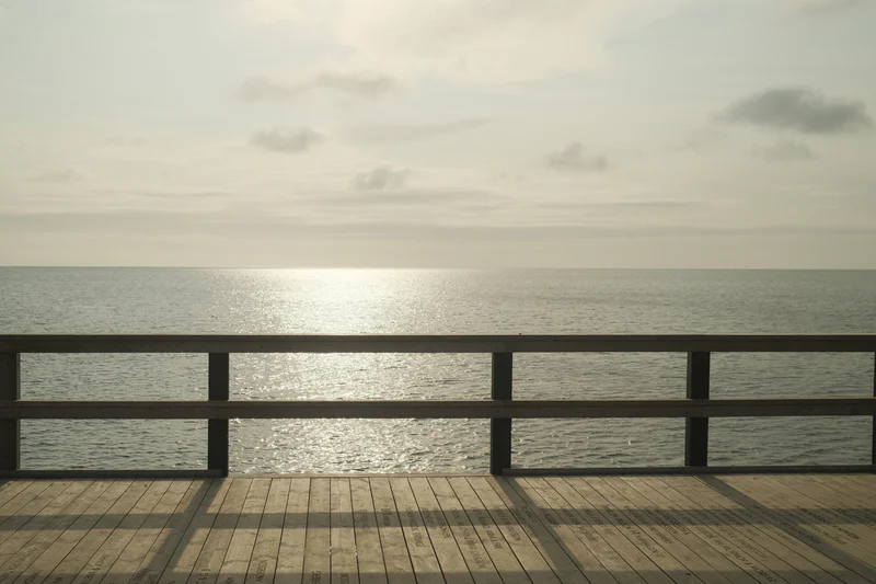 Wooden deck with a railing overlooking calm ocean and a low sun near the horizon under cloudy sky.