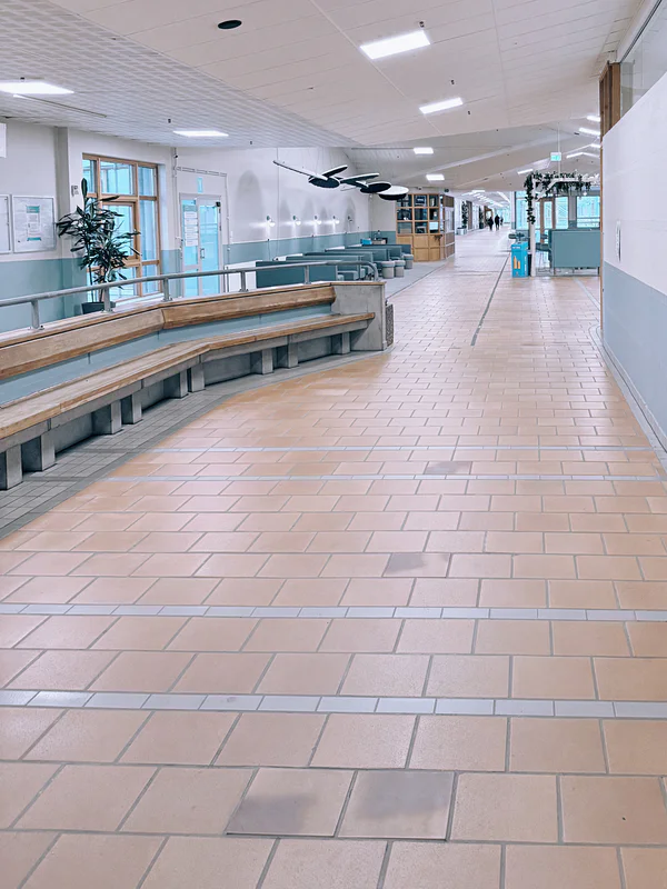 Bright school corridor with tiled floor, benches, windows, and ceiling lights stretching into the distance.