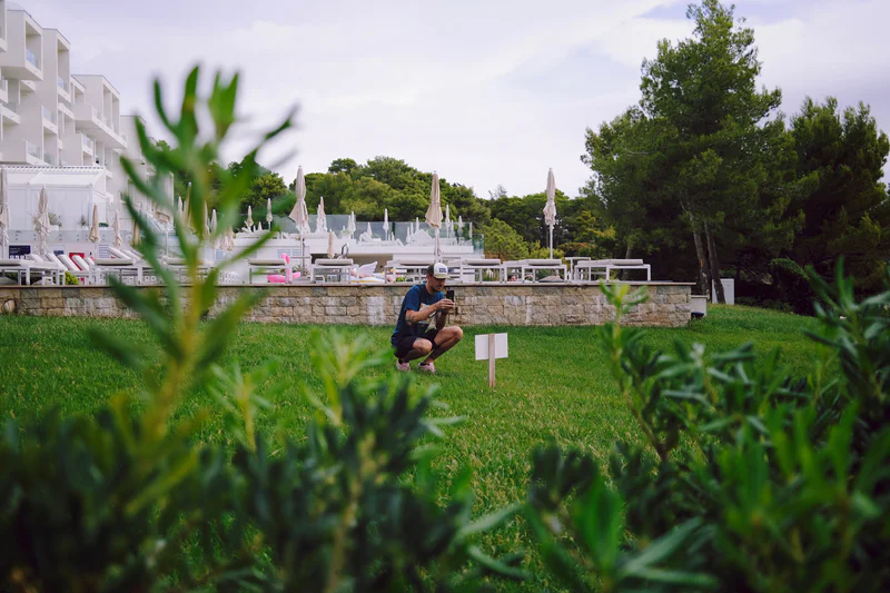 Man kneels on a lawn beside a white marker, with hotel buildings and umbrellas in the background.