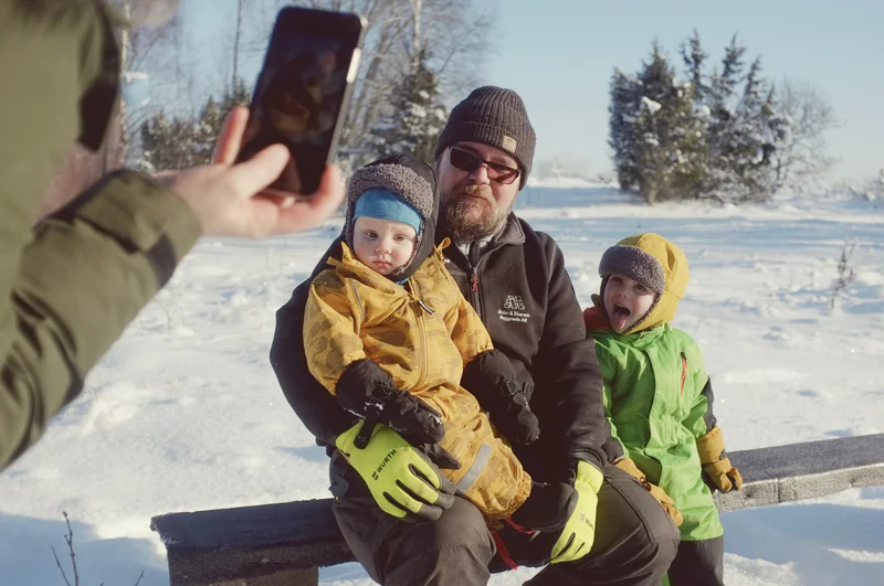 Man in winter clothing holds a bundled toddler while two others sit outdoors in a snowy landscape.