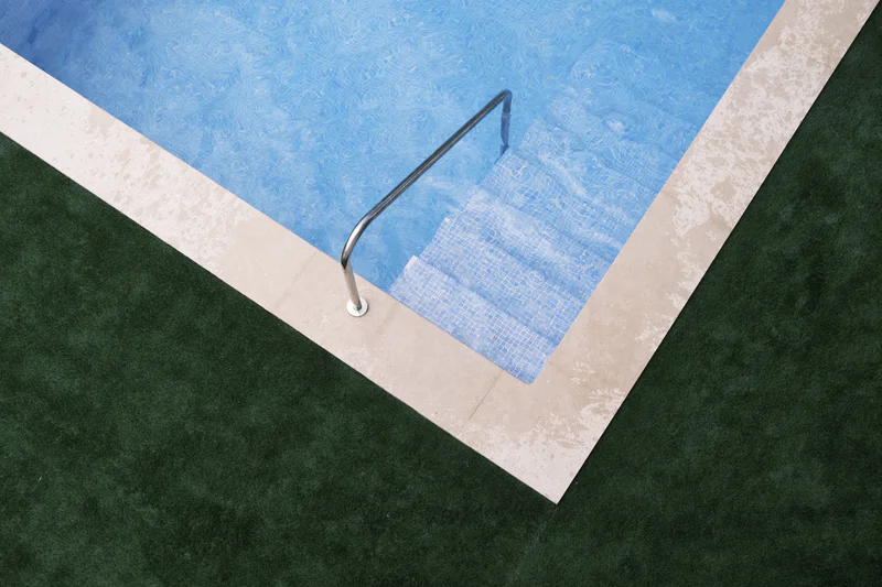 Rectangular swimming pool filled with blue water, bordered by white concrete and green turf.