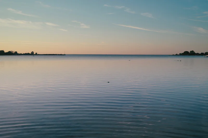 Calm lake at dusk with rippling water, distant shoreline, and a pink-blue sky.