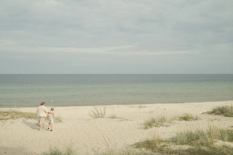 Two people walking on sandy dunes beside a calm ocean under an overcast sky.