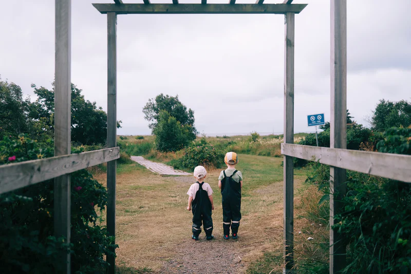 Two children in caps walk down a gravel path between wooden gate posts outdoors.