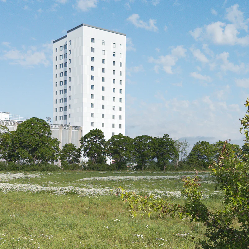Tall white apartment building with many windows behind trees under a partly cloudy blue sky.