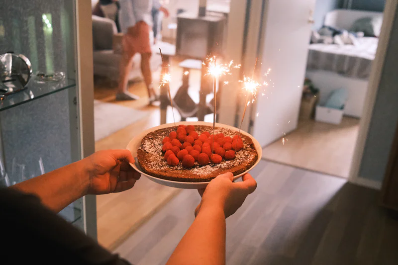 Person holds a round cake with glowing sparklers and red berries on a tray indoors.