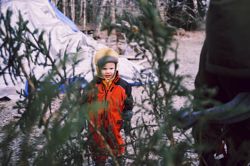 Child in an orange winter jacket and hat stands outdoors in snow behind branches and a tent.