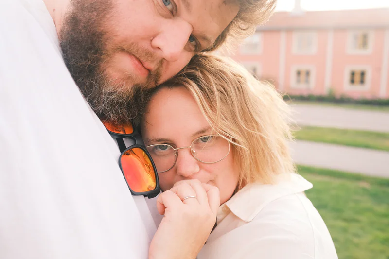 Man and woman lean together outdoors, the woman wearing glasses and resting her hand near her face.