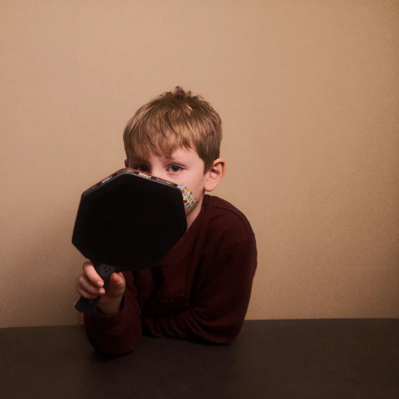 Child holding an octagonal black table tennis racket toward the camera against a plain wall.