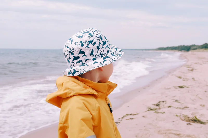 Child in a yellow raincoat and patterned bucket hat stands on a sandy beach by the ocean.