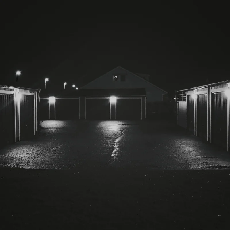 Night view of garage doors and fencing with bright lights along a driveway.
