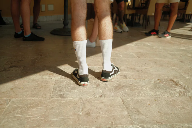 Group standing indoors wearing white socks and dark sneakers, legs visible in foreground on a tiled floor.