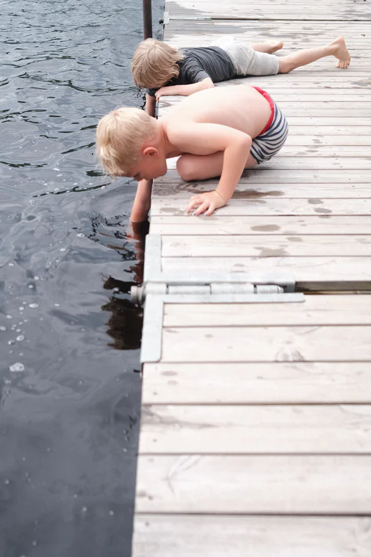 Two shirtless boys crawl on a wooden dock edge above the water, with one reaching forward while bending.
