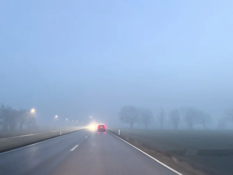 Misty highway at dusk with a car ahead and headlights fading into fog.