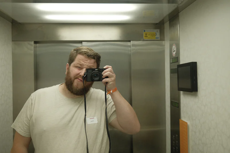 Bearded man taking a mirror selfie with a camera inside an elevator.