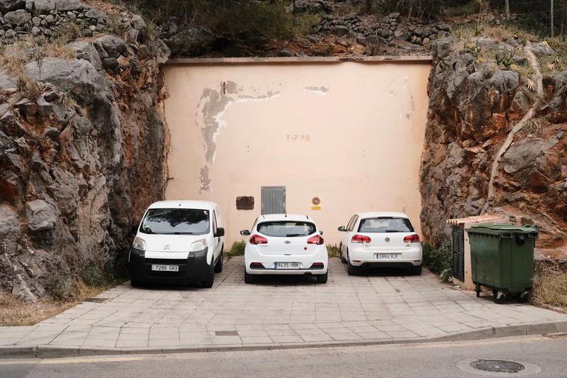 Three parked white cars in a rocky roadside cutout, with a green waste bin on the right.