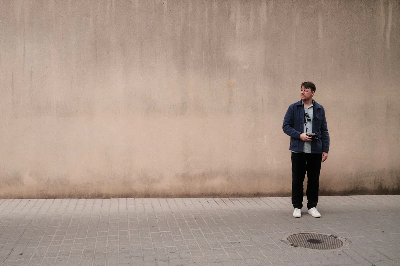 Man in a blue jacket stands on a paved walkway beside a blank concrete wall, holding a camera.