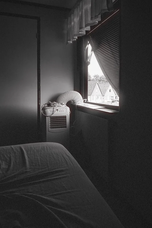 Portable air conditioner beside a window with blinds, with part of a bed in the foreground.