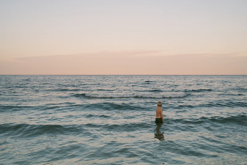 Person standing waist-deep in ocean waves at sunset, with calm horizon and pastel sky.
