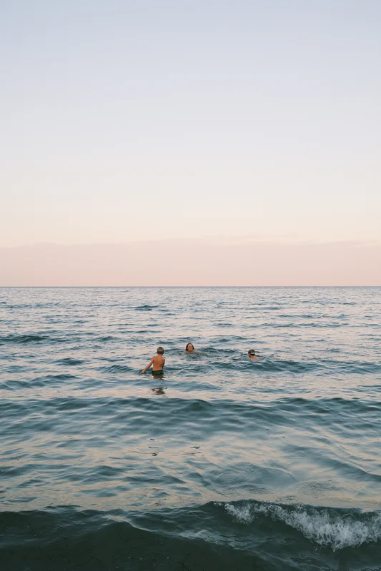 Three swimmers stand chest-deep in calm ocean water at sunset under a pale sky.