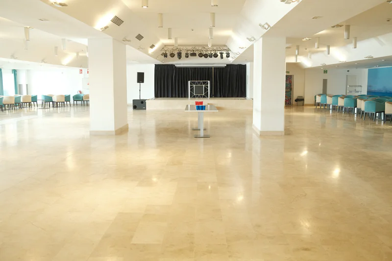 Empty indoor hall with stage curtains, speakers, lighting rig, and rows of blue chairs along the sides.