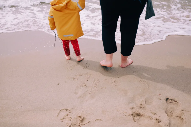 Child in yellow raincoat and red leggings stands near an adult on a sandy beach, barefoot by waves.