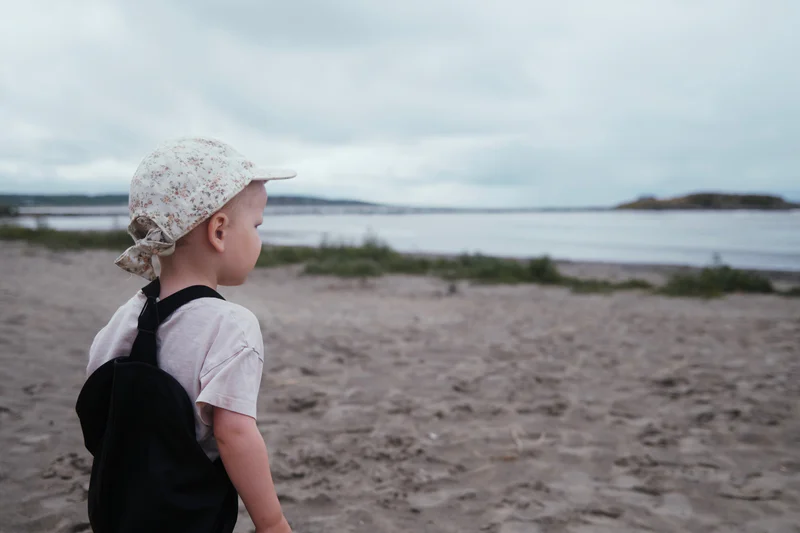 Child wearing a floral sun hat and black backpack stands on a sandy beach facing the water.
