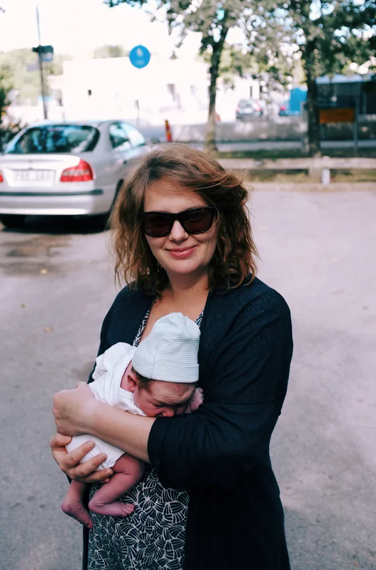 Woman wearing sunglasses holds a baby wrapped in a blanket outdoors in a parking lot.