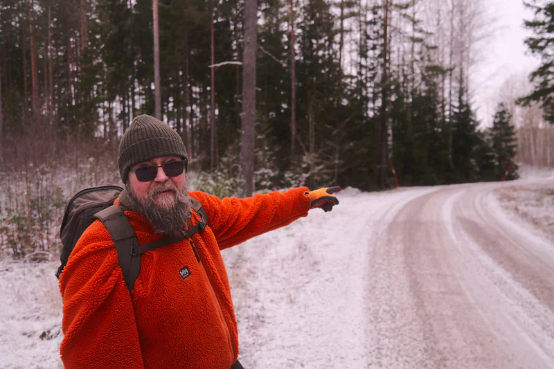 Bearded man in an orange winter coat and knit hat pointing along a snowy forest road.