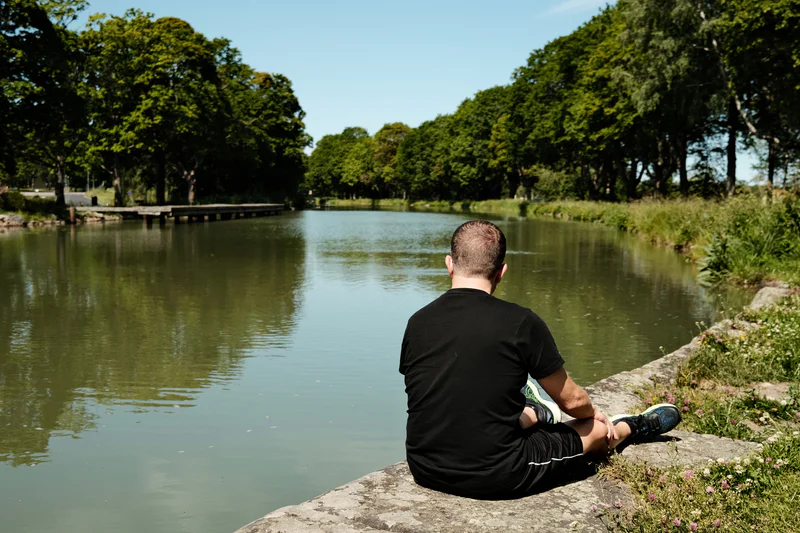 Man sitting on a riverside rock beside still green water, lined with trees under a blue sky.