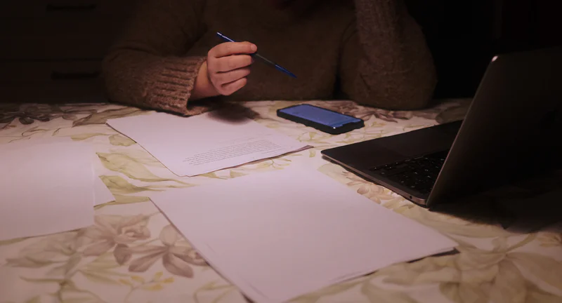 Person writing with a blue pen at a desk covered with papers, a phone, and a laptop.