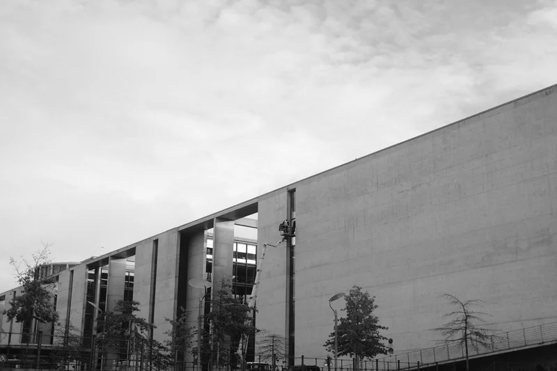 Modern concrete building facade with large overhanging roof, trees, and cloudy sky.