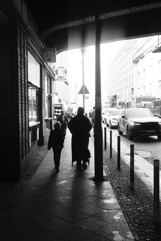 Black-and-white city sidewalk with pedestrians walking past parked cars under an arched storefront canopy.
