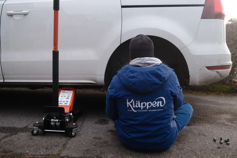 Person in blue jacket kneeling near a jack beside a white vehicle with a wheel removed
