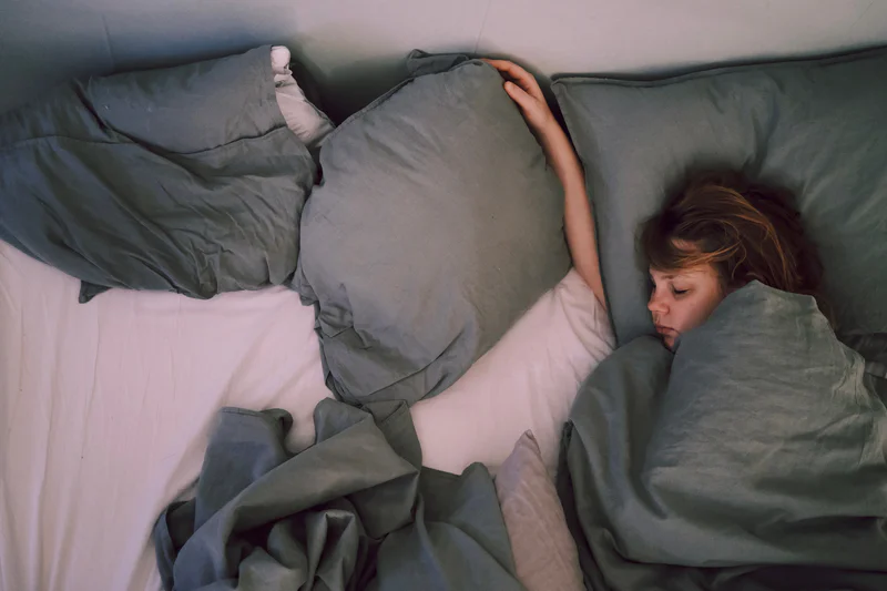 Woman lying on a bed under gray covers, holding a blanket above her face while looking down.