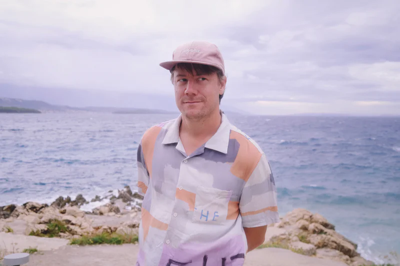 Man wearing a pink cap and striped shirt standing by a rocky shoreline with the ocean behind him.