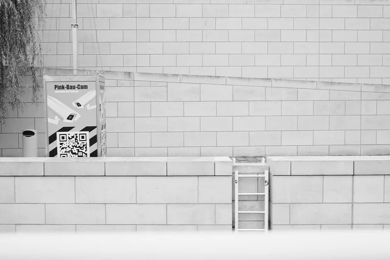Black and white wall with a small poster showing “Pink-Bau-Cam” and a QR code, plus a ladder.