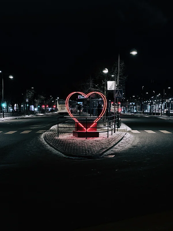 Night street intersection with red illuminated heart sculpture and crosswalk lines under streetlights.