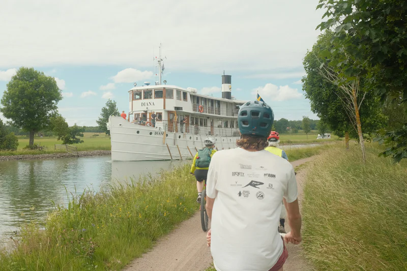 A man on a riverside path bikes toward a canal boat, with several cyclists and trees nearby.