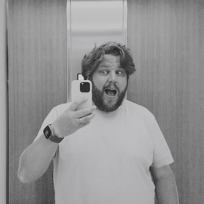 Black-and-white mirror selfie of a bearded man taking a phone picture in a doorway.