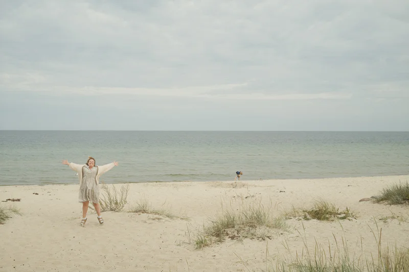Person in a light dress runs on a sandy beach with ocean and cloudy sky behind.