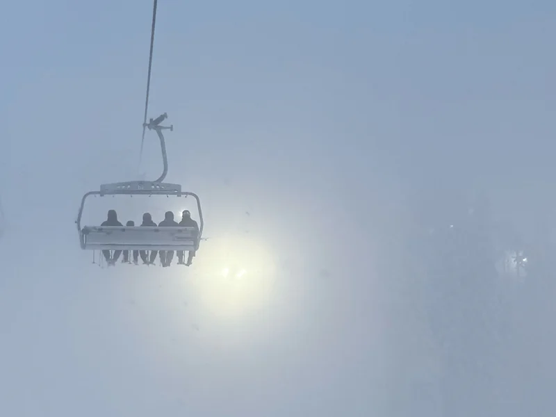 Snow-covered sky with a cable car gondola hanging in light haze, with sunlight behind it.