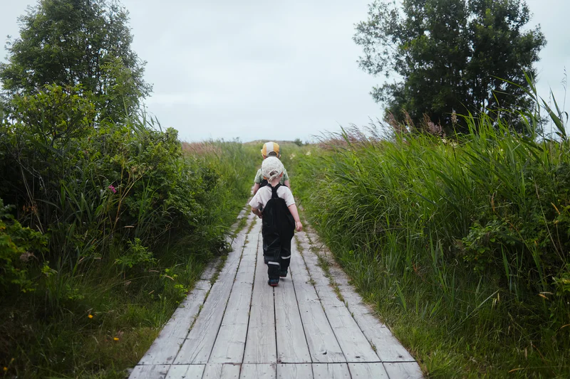 Two children walk on a narrow wooden path through tall grass and trees.