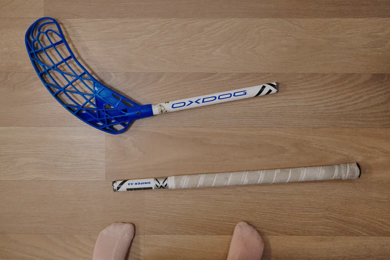 Blue and white floorball stick lies on a wooden floor, with the stick broken into two pieces.