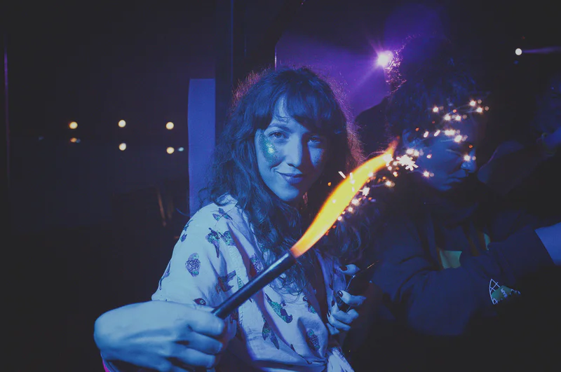 Woman in a party setting holds a flaming sparkler stick producing sparks toward the camera.